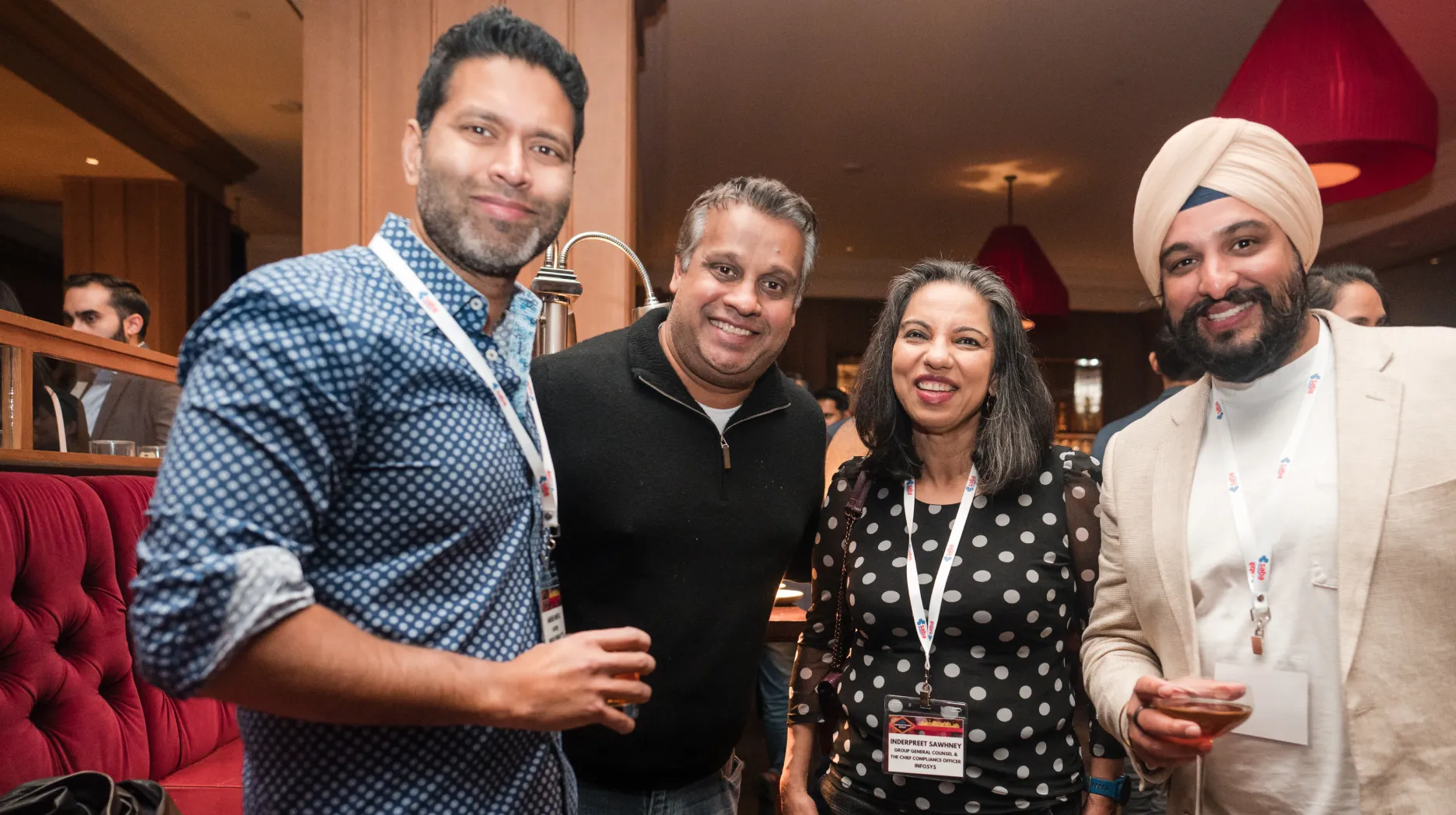 Four smiling people posing at an indoor event, two men, one woman with a name badge, and one man wearing a beige turban, all holding drinks.